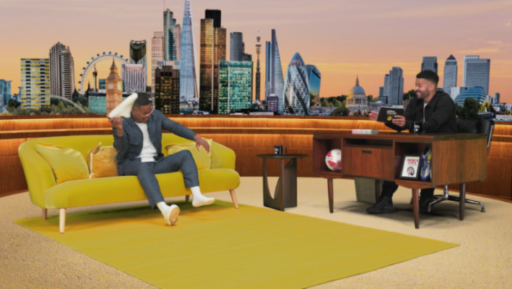 Two men in a studio with a London skyline backdrop, one on a yellow couch holding a shoe, the other at a desk.