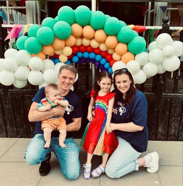 A family of four kneeling in front of a rainbow balloon arch.