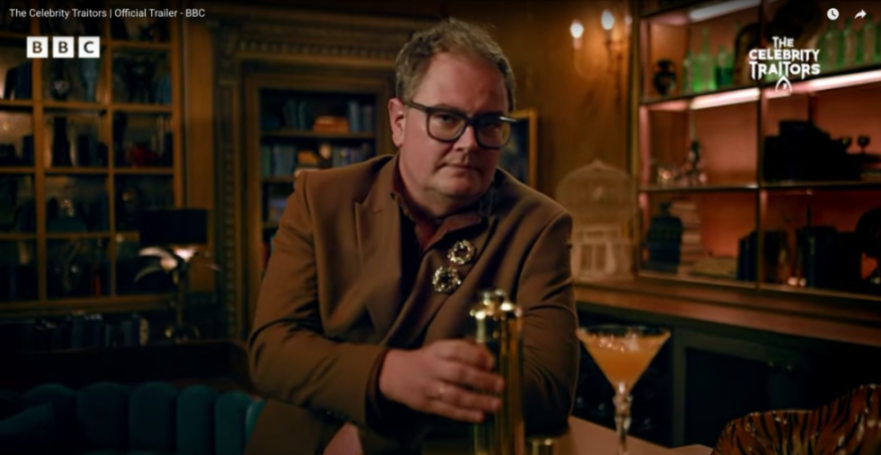 Alan Carr in a brown suit and glasses holding a cocktail shaker.
