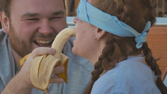 Man feeding blindfolded woman a banana.