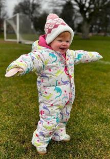 Toddler in dinosaur-print snowsuit on grassy field.