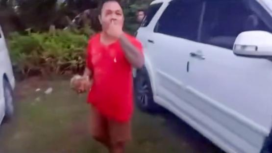 A man in a red shirt holding something small, possibly near a car.