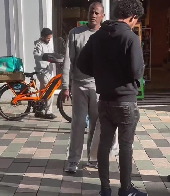 Three men on a paved area, one with an orange bicycle with a delivery bag.