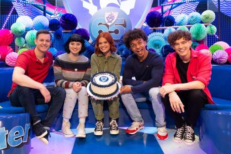 Five presenters on a children's television program celebrating its 65th birthday with a cake.