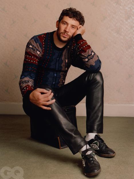 Josh O'Connor in a multi-colored sweater, black leather pants, white socks, and black and white athletic shoes, sitting on a black suitcase.