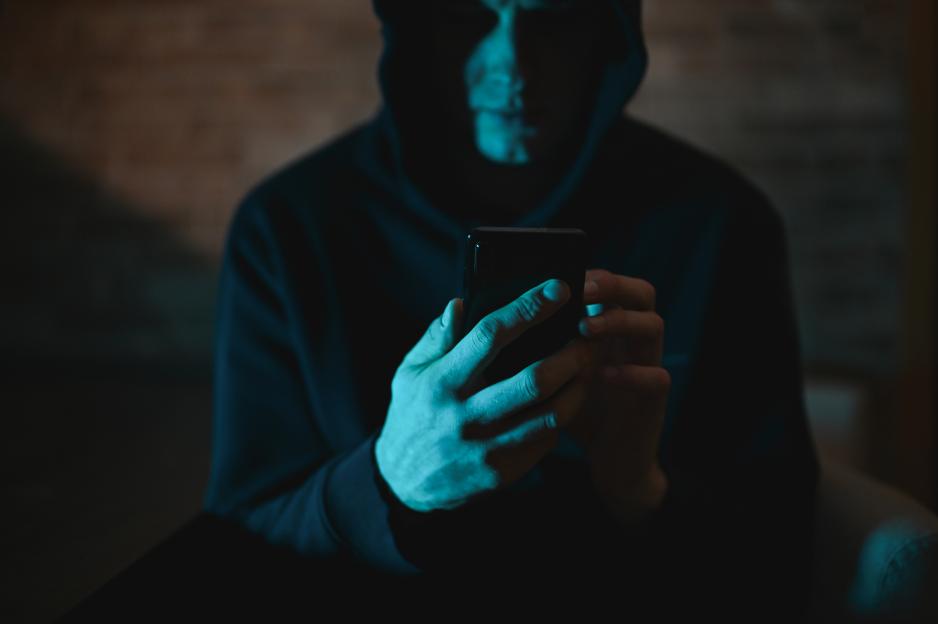 A man in a hooded sweatshirt typing on a laptop, silhouetted against a dark background.
