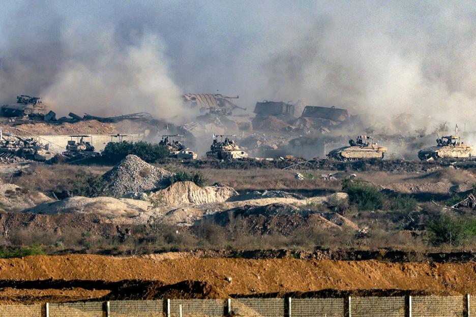 Israeli army vehicles deployed across the border fence in the Gaza Strip, viewed from southern Israel, with smoke and debris in the background.