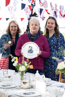 Three women at a table displaying a commemorative plate honoring Arnold Sharrocks, a WWII hero.