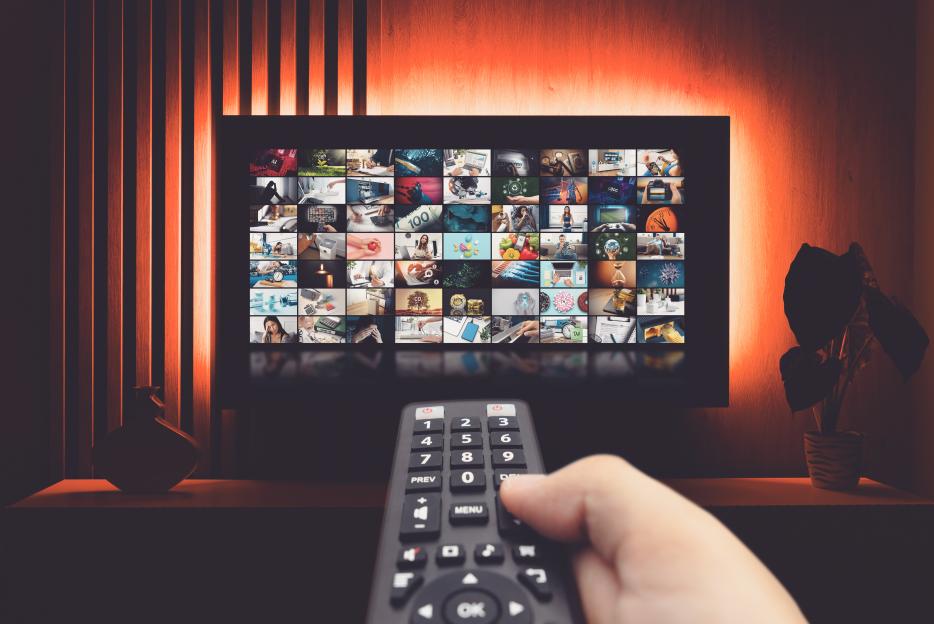 Hand holding a remote control pointed at a TV screen displaying a collage of multimedia streaming content.