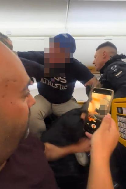 A man in a black sweatshirt and blue hat being apprehended by a police officer on a plane while another man records the scene on his phone.