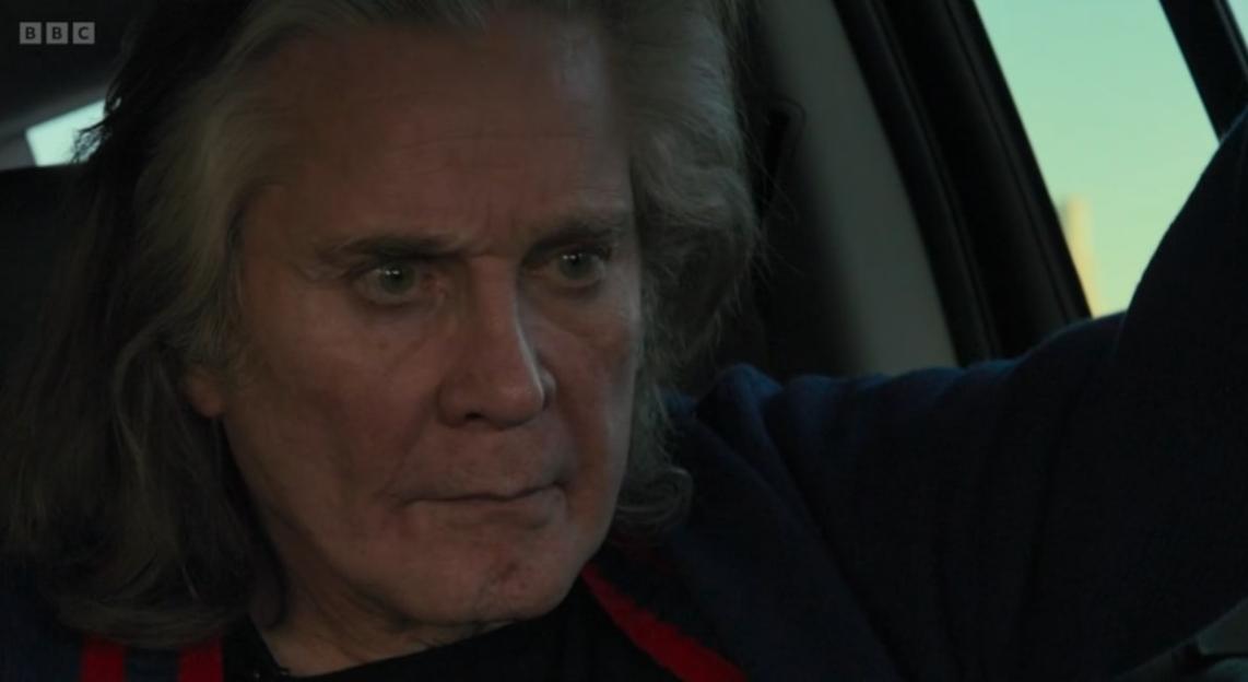 Ozzy Osbourne with long hair and a serious expression, sitting in a car.
