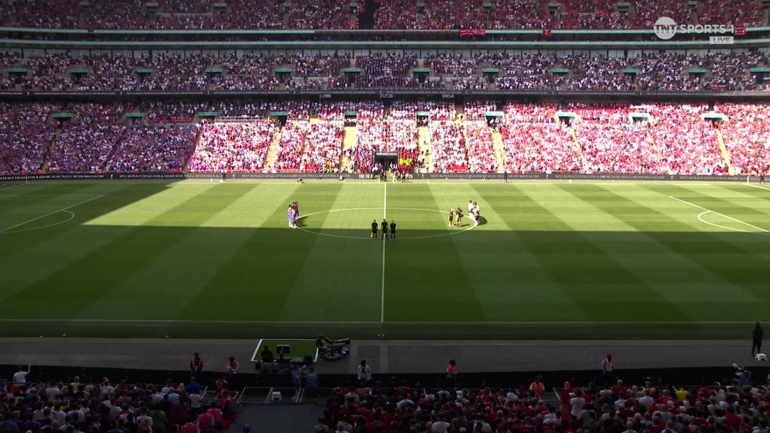 Soccer players standing on the field before a large crowd in a stadium.