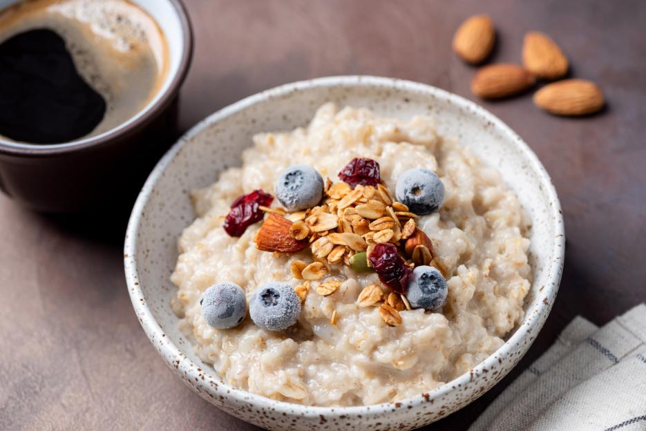 Oatmeal porridge bowl with frozen blueberries, granola, almonds, and dried cranberries, with a cup of coffee and almonds in the background.