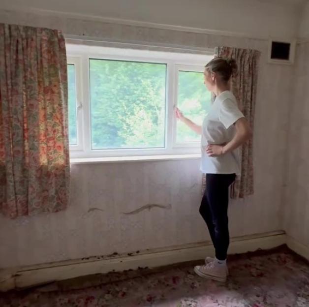 A woman opening a window in a dilapidated room with floral wallpaper and carpet.