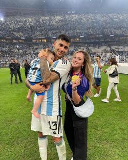 A man in an Argentinian soccer jersey holds a baby while standing next to a woman holding a gold medal; they are in a stadium.