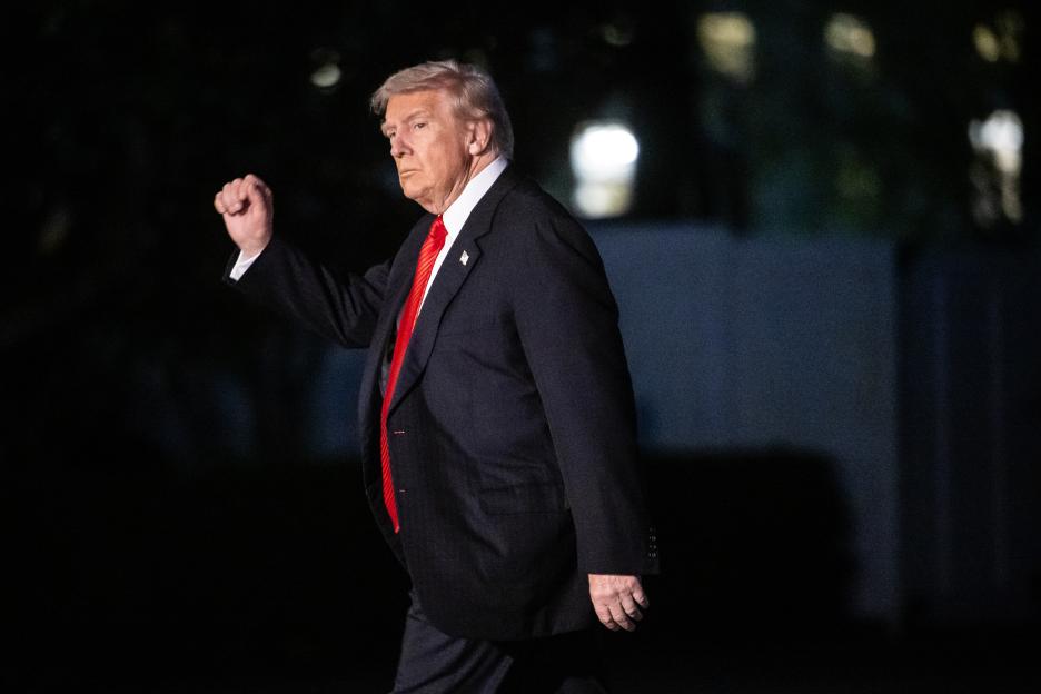 Donald Trump, wearing a red tie, with a clenched fist, walks across the South Lawn of the White House at night.