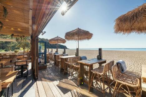Beachfront restaurant patio with wooden tables and wicker chairs under thatched umbrellas.