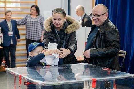 A young boy drops a ballot into a transparent ballot box while being assisted by two adults at a polling station.
