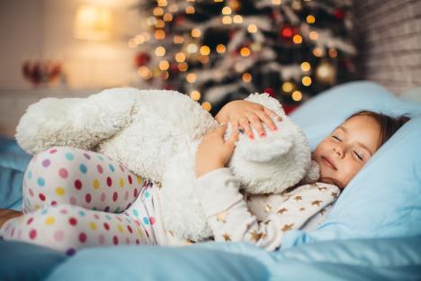 Girl sleeping in bed with teddy bear, Christmas tree in background.