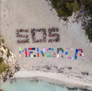 Aerial view of protestors on a beach forming the letters SOS MENORCA with their bodies and towels.