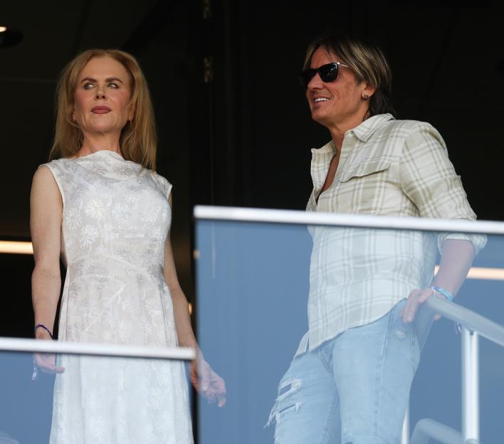 Nicole Kidman in a white dress and Keith Urban in a plaid shirt and sunglasses watching a soccer game.
