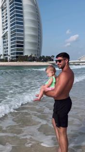 Man holding baby at the beach with a tall building in the background.