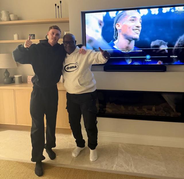 Cole Palmer posing with a friend in front of a TV showing a football match.