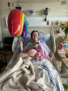 Woman in hospital bed holding rainbow balloon, surrounded by gifts.