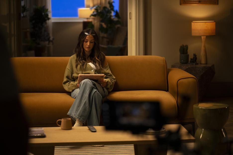 Michelle Keegan sits on an orange couch in a living room, looking at a tablet.