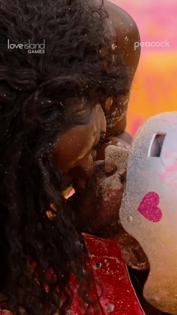Two people covered in sand kissing, one wearing a helmet with a pink heart.