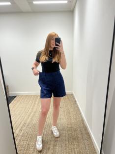 Woman in dark top and denim shorts taking a selfie in a mirror.