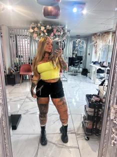A tattooed woman in a yellow crop top and black shorts takes a selfie in a hair salon.