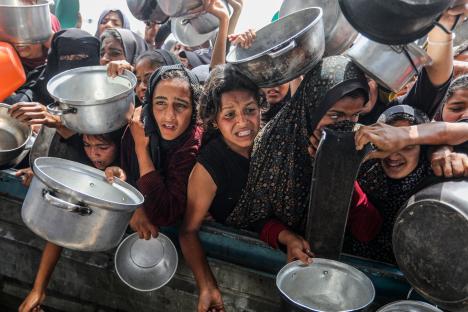 Palestinian women and children holding pots and bowls during a food distribution in Khan Yunis, Gaza.