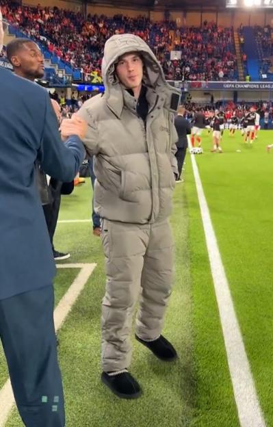 A man in a padded gray jacket and pants, wearing black slippers, stands on a soccer field, shaking hands with another man.
