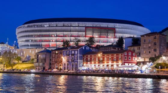 Night view of the San Mames Stadium in Bilbao, Spain, with buildings and a river in the foreground.