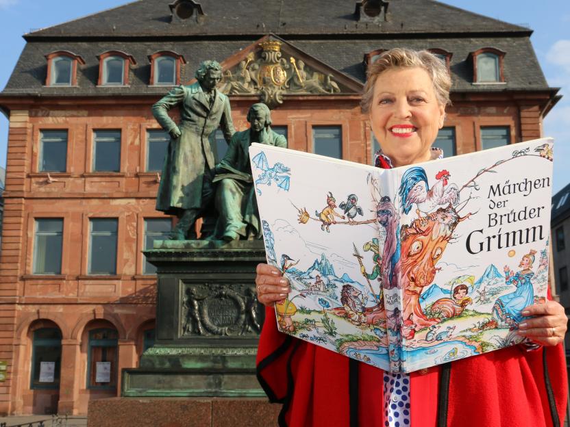 Woman holding a large illustrated book of Grimm's fairy tales in front of a statue of the Brothers Grimm.