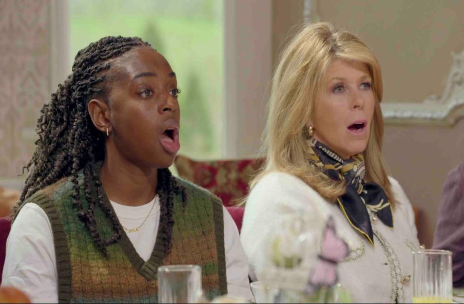 Two women, one Black with braided hair in a green sweater vest, and one white with blonde hair in a white sweater, looking surprised with their mouths agape.