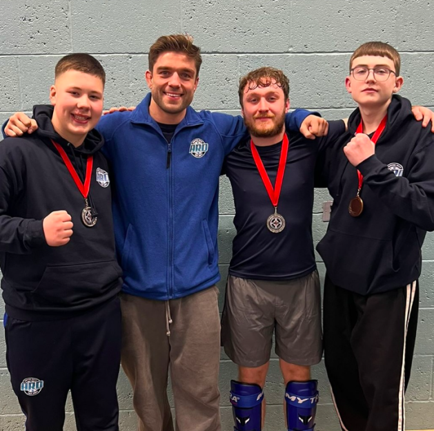 Four young men with medals around their necks stand with their arms around each other.