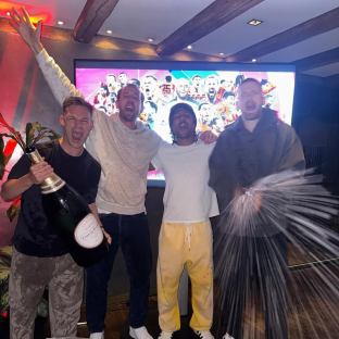 Four men celebrating with champagne in front of a large screen showing a collage of soccer players.