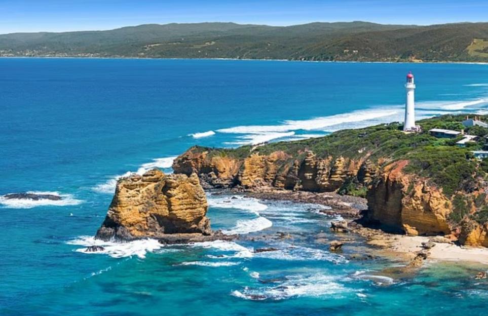 An aerial view of a white lighthouse with a red roof on a green cliff overlooking a blue ocean with waves crashing on rock formations and a sandy beach.