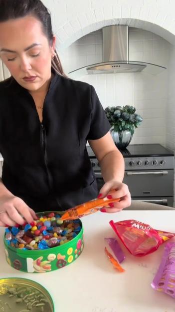 A person filling a green tin with various candies, including jelly beans and wrapped sweets.