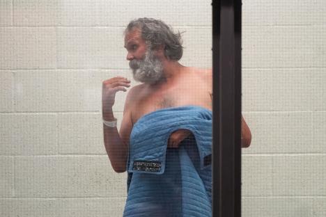 Shirtless man with long gray hair and beard seen through glass, holding a blue blanket.