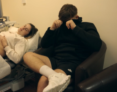 A pregnant woman lies on an examination table while a man sits nearby, covering his face with his hands.