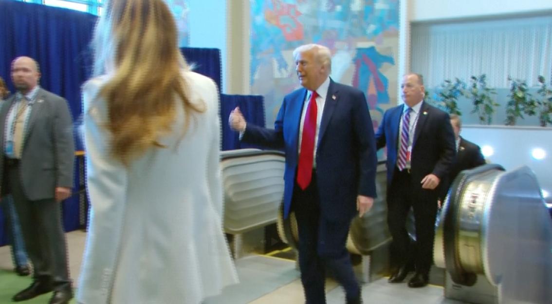 Donald Trump walking up a broken escalator, with a woman and security guards in front and behind him.