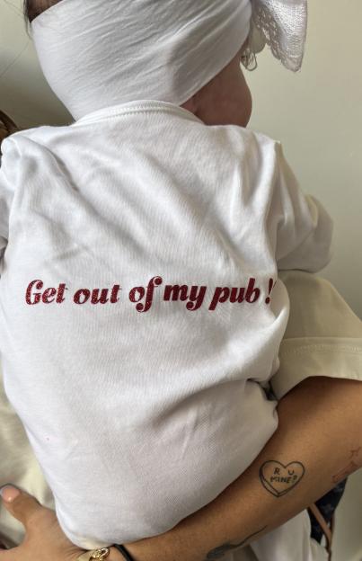 A baby wearing a white shirt with red glitter text that reads "Get out of my pub!"
