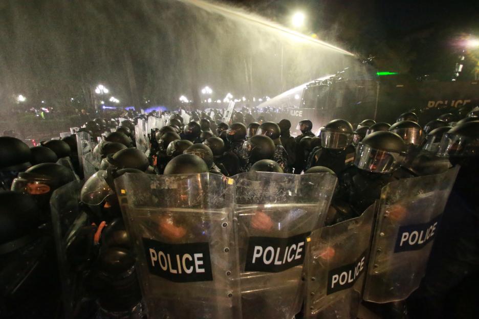 Police in riot gear with shields forming a line, with a water cannon spraying water from behind them.