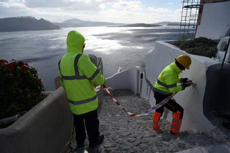 Two workers in bright yellow safety vests repair a building overlooking a body of water.