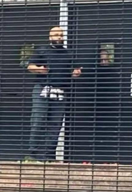 A man in dark clothing, possibly the alleged terrorist, stands behind a security gate.