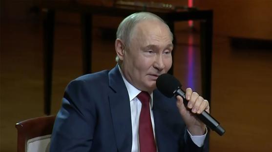 Vladimir Putin speaking into a microphone, stating that love is shown in ways beyond kissing and hugging.