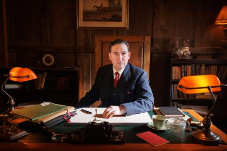 A man in a suit sits at a desk in a study, reviewing papers.